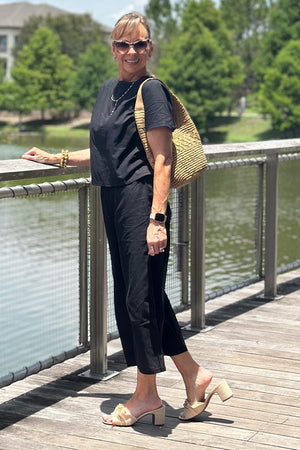 Woman wearing black loose fit co-ord set with short sleeves and wide-leg pants, accessorized with sunglasses and a woven tote bag on a wooden boardwalk by the water.