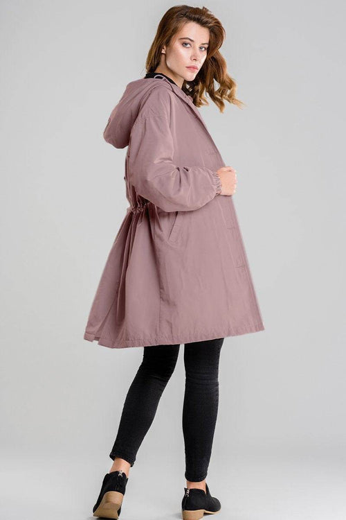 Woman wearing pink water resistant oversized hooded windbreaker jacket with adjustable waist and long length over black pants and black shoes.