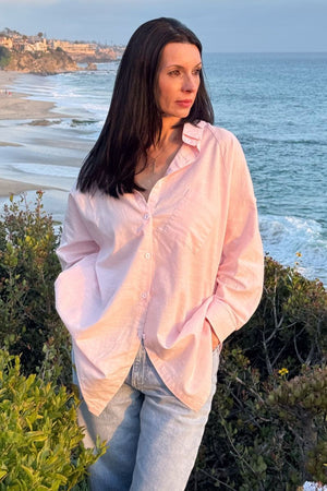 Woman wearing oversized linen shirt in sun-washed pink by the ocean coastline