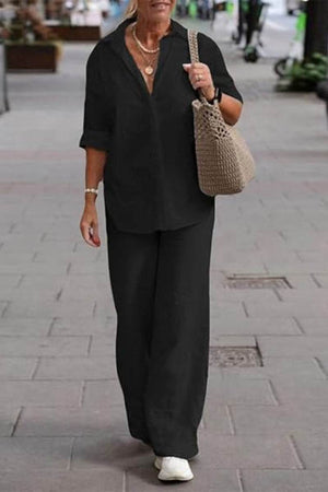 Woman wearing oversized 100% cotton black shirt and wide-leg pants co-ord set walking outdoors with a beige woven shoulder bag and white sneakers.
