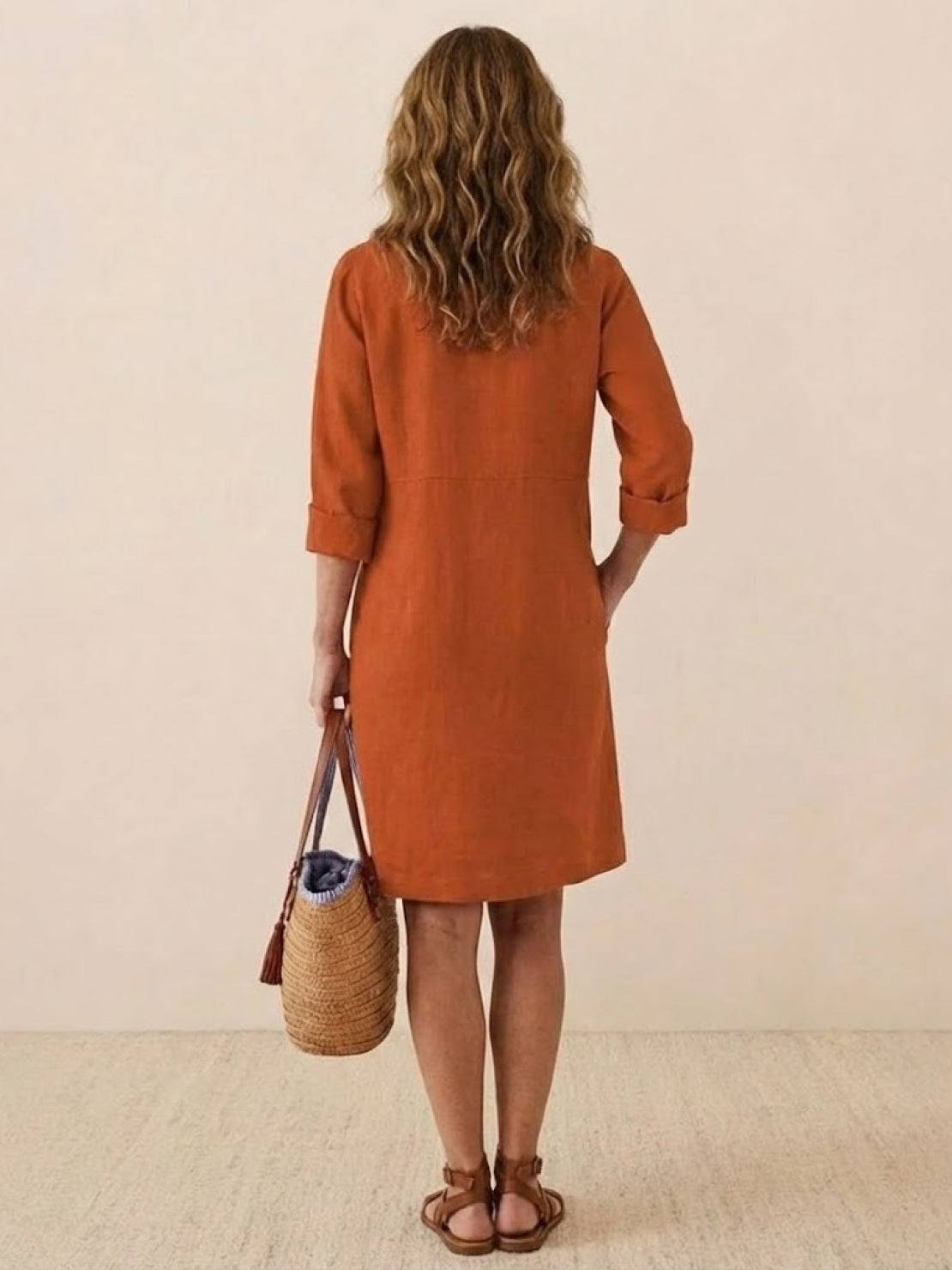 Woman wearing rust-colored rolled sleeve cotton dress carrying straw handbag viewed from back