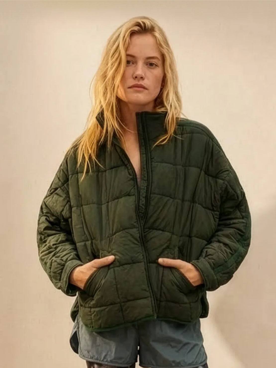 Woman wearing dark green lightweight puffer travel jacket with hands in pockets against neutral background