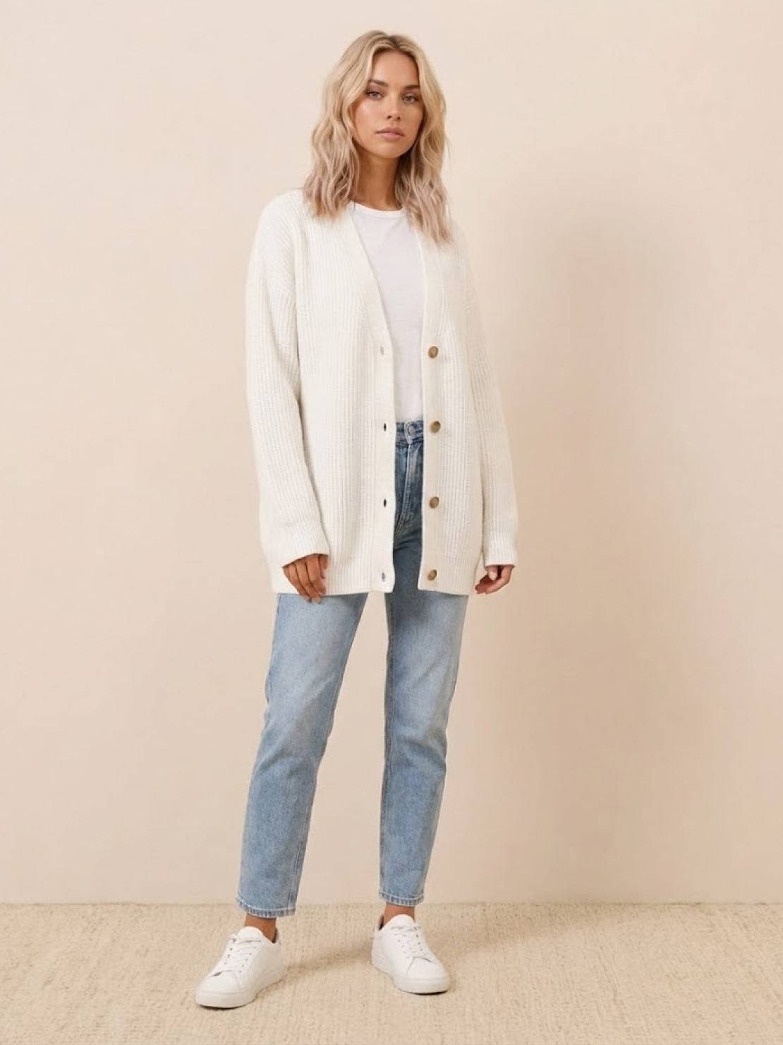 woman wearing relaxed fit oversized white button down knitwear cardigan with jeans and white sneakers indoors
