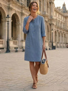 Woman wearing casual blue cotton dress with 3/4 sleeves and front buttons walking outdoors carrying a straw handbag.
