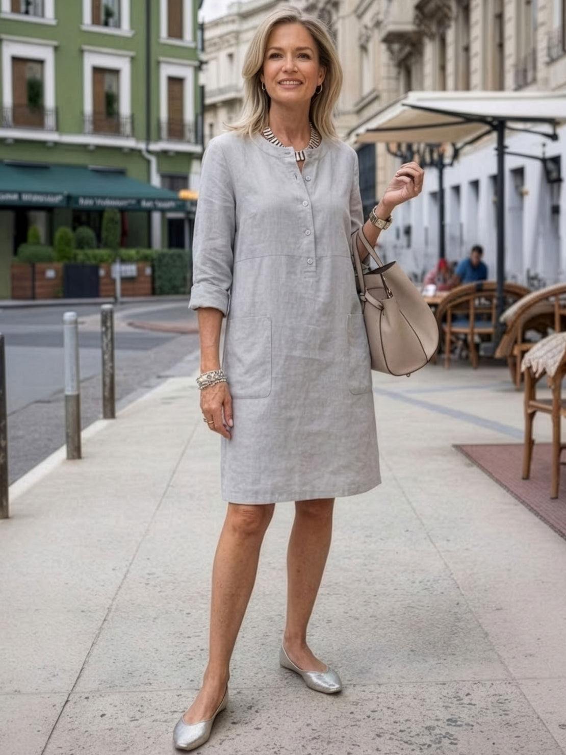 woman wearing gray cotton dress with rolled-up sleeves standing outdoors in city street with buildings and café seating in background