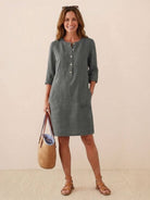 woman wearing gray knee-length cotton dress with 3/4 rolled sleeves and button front carrying straw tote bag and sandals