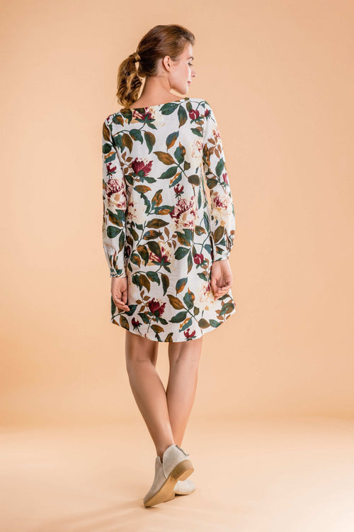 Woman wearing an all over floral print linen shift dress with long sleeves and beige shoes against a peach background.